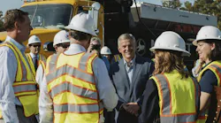 North Carolina Gov. Josh Stein smiles while talking with a group of people in hard hats and high-visibility safety vests, with a yellow dump truck parked behind them.