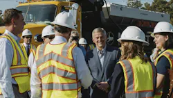 North Carolina Gov. Josh Stein smiles while talking with a group of people in hard hats and high-visibility safety vests, with a yellow dump truck parked behind them.