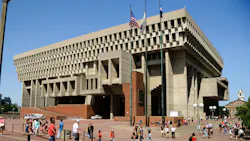 Exterior view of Boston City Hall, a large concrete Brutalist building, with pedestrians crossing the brick plaza in front and flags flying above the entrance.