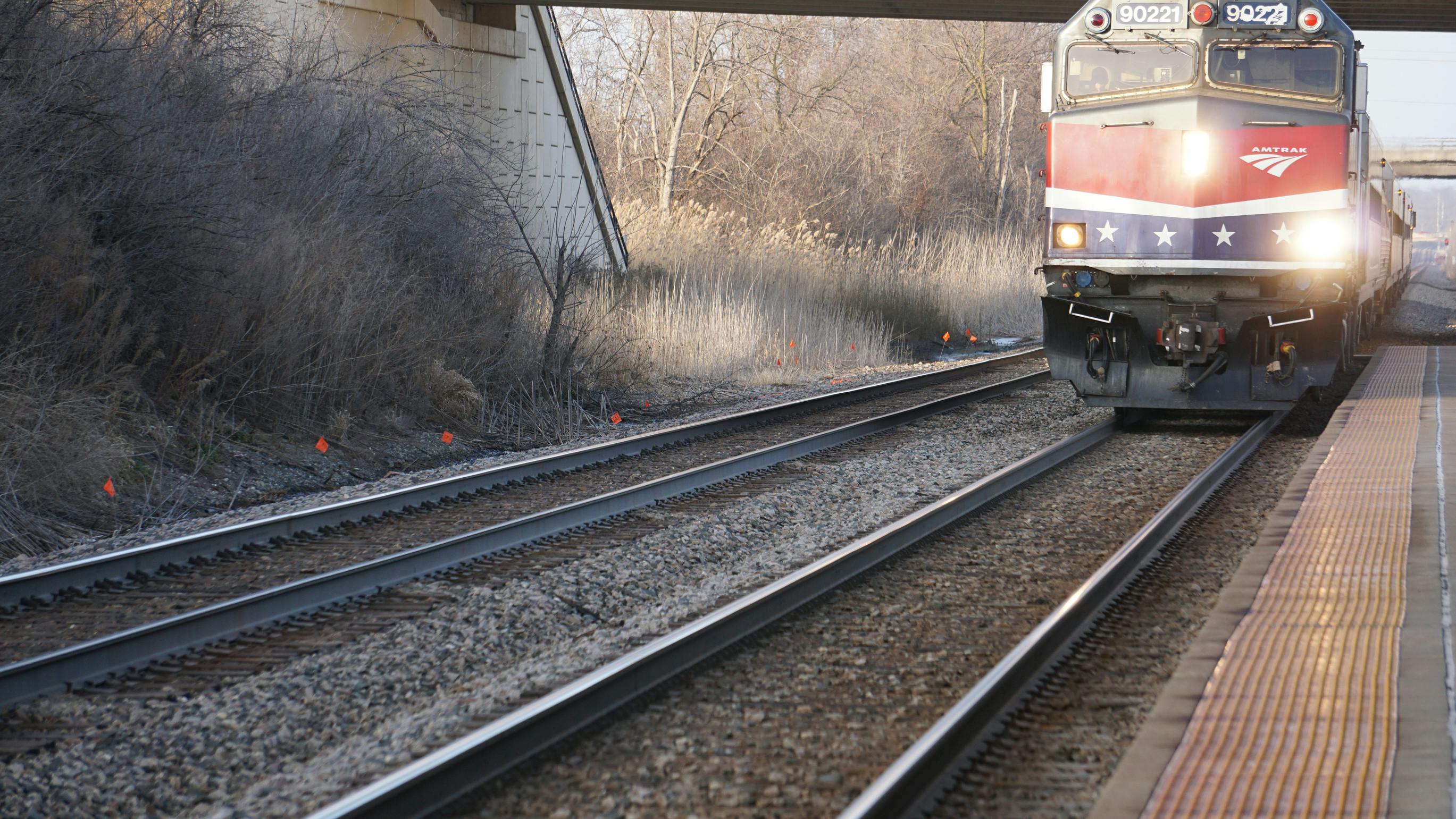An Amtrak locomotive, unit 90221, in red, white and blue livery with white stars approaches a station platform with its headlights on, running beneath an overpass beside multiple tracks and bare winter vegetation.