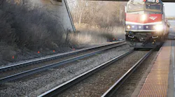 An Amtrak locomotive, unit 90221, in red, white and blue livery with white stars approaches a station platform with its headlights on, running beneath an overpass beside multiple tracks and bare winter vegetation.