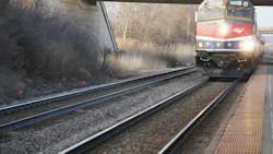 An Amtrak locomotive, unit 90221, in red, white and blue livery with white stars approaches a station platform with its headlights on, running beneath an overpass beside multiple tracks and bare winter vegetation.