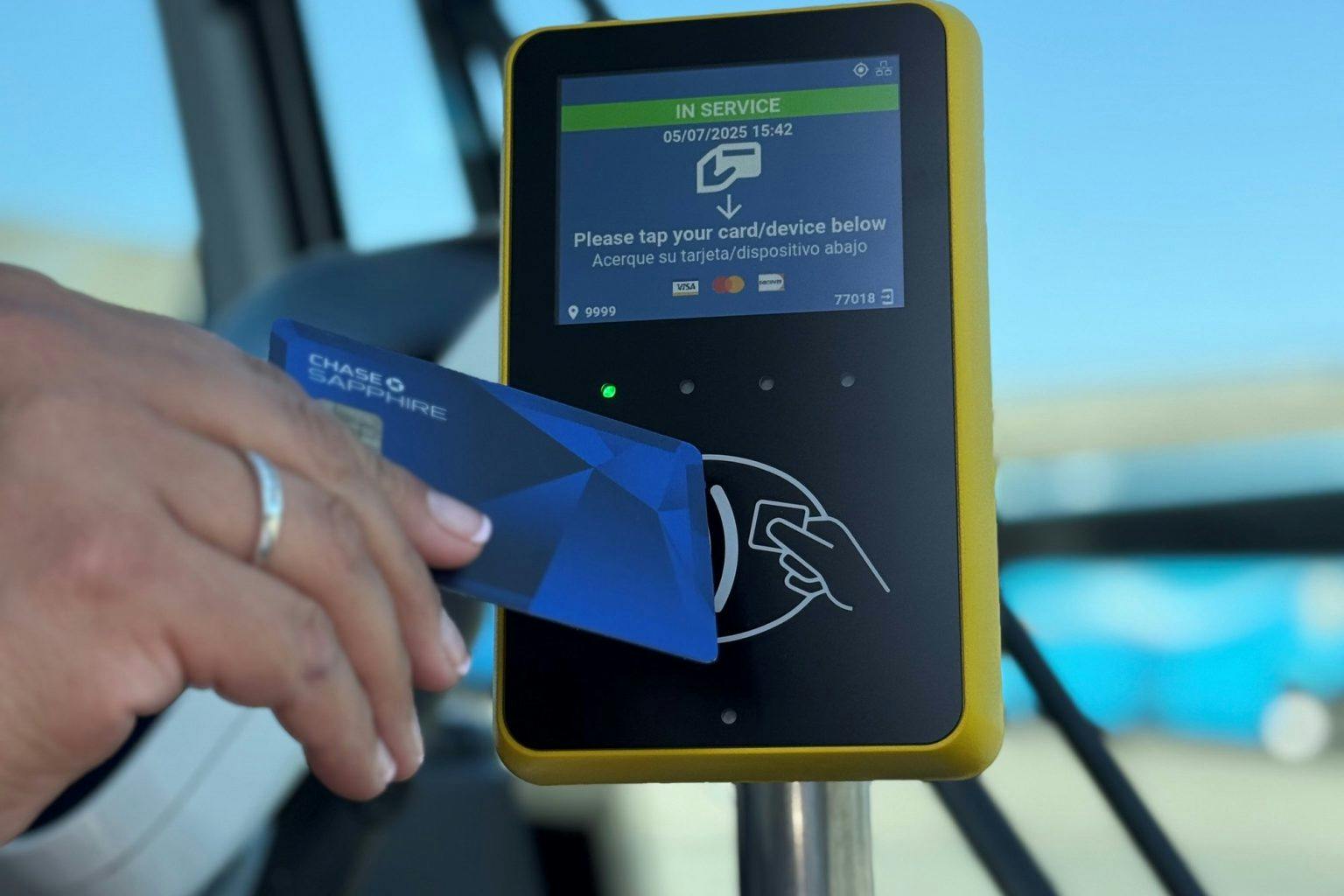 A hand taps a Chase Sapphire credit card on a yellow open-payment fare reader displaying &lsquo;In Service, Please tap your card/device below.
