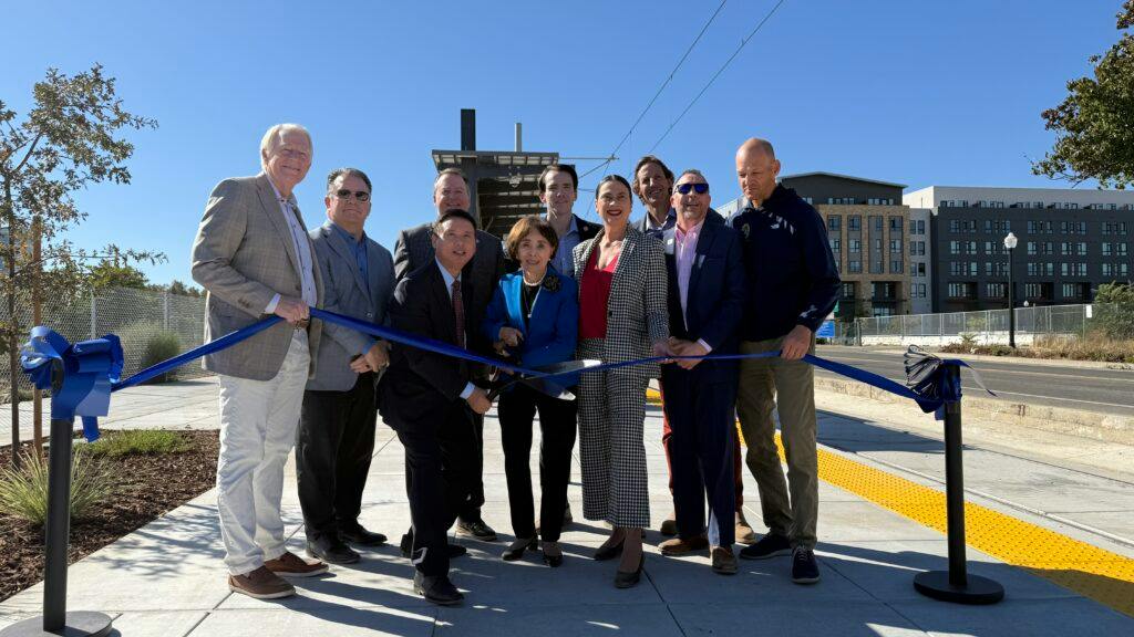 A group of nine officials and community members holds a blue ribbon with ceremonial scissors on a paved platform beside a yellow tactile warning strip, with overhead wires, a small station shelter and apartment buildings behind them.