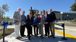 A group of nine officials and community members holds a blue ribbon with ceremonial scissors on a paved platform beside a yellow tactile warning strip, with overhead wires, a small station shelter and apartment buildings behind them.