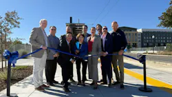 A group of nine officials and community members holds a blue ribbon with ceremonial scissors on a paved platform beside a yellow tactile warning strip, with overhead wires, a small station shelter and apartment buildings behind them.