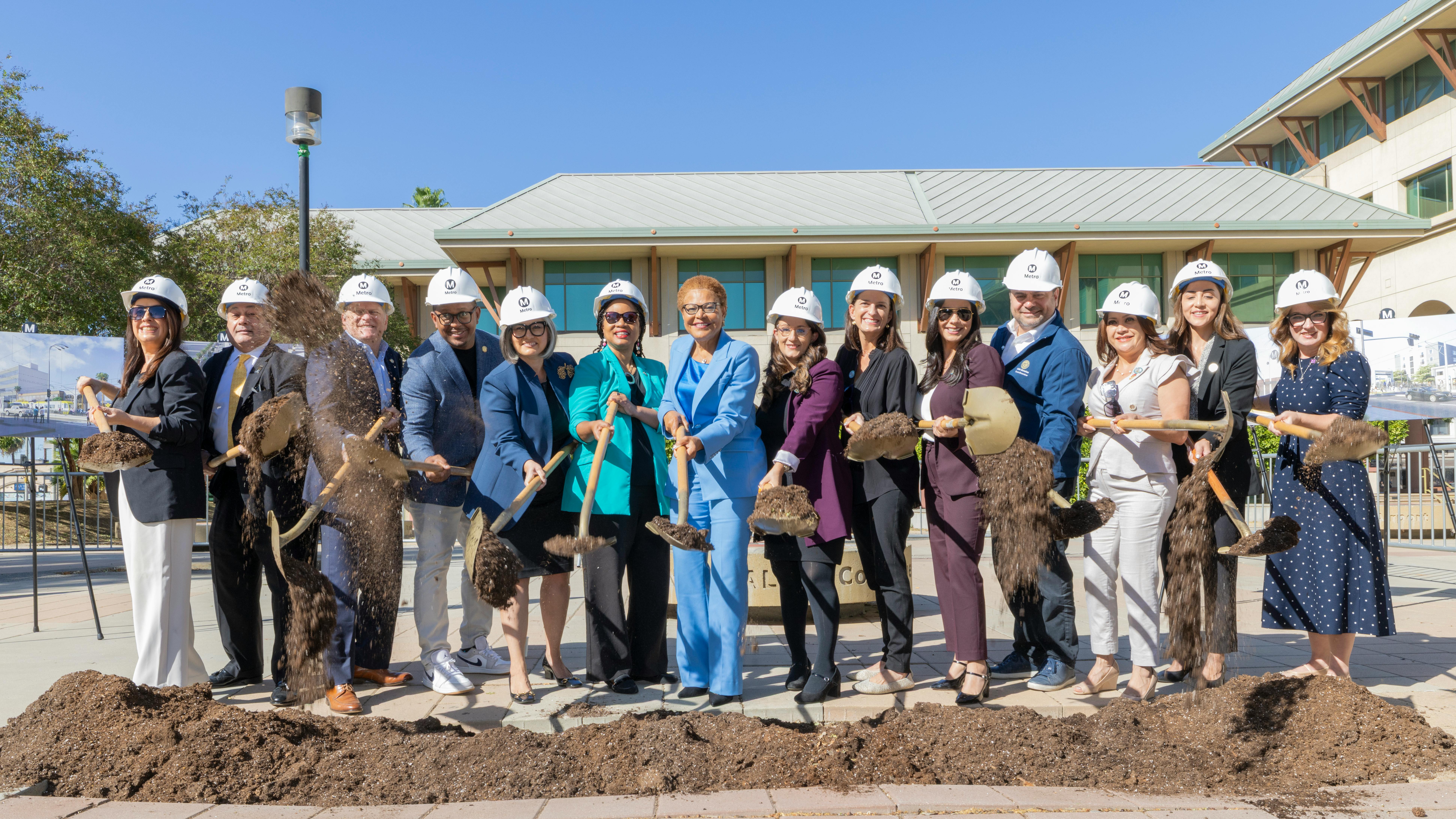 A row of fourteen people in white LA Metro hard hats take part in a groundbreaking ceremony, tossing dirt with gold shovels outside a building.