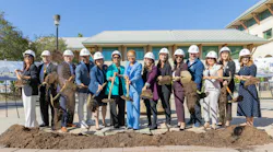 A row of fourteen people in white LA Metro hard hats take part in a groundbreaking ceremony, tossing dirt with gold shovels outside a building.