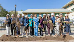 A row of fourteen people in white LA Metro hard hats take part in a groundbreaking ceremony, tossing dirt with gold shovels outside a building.