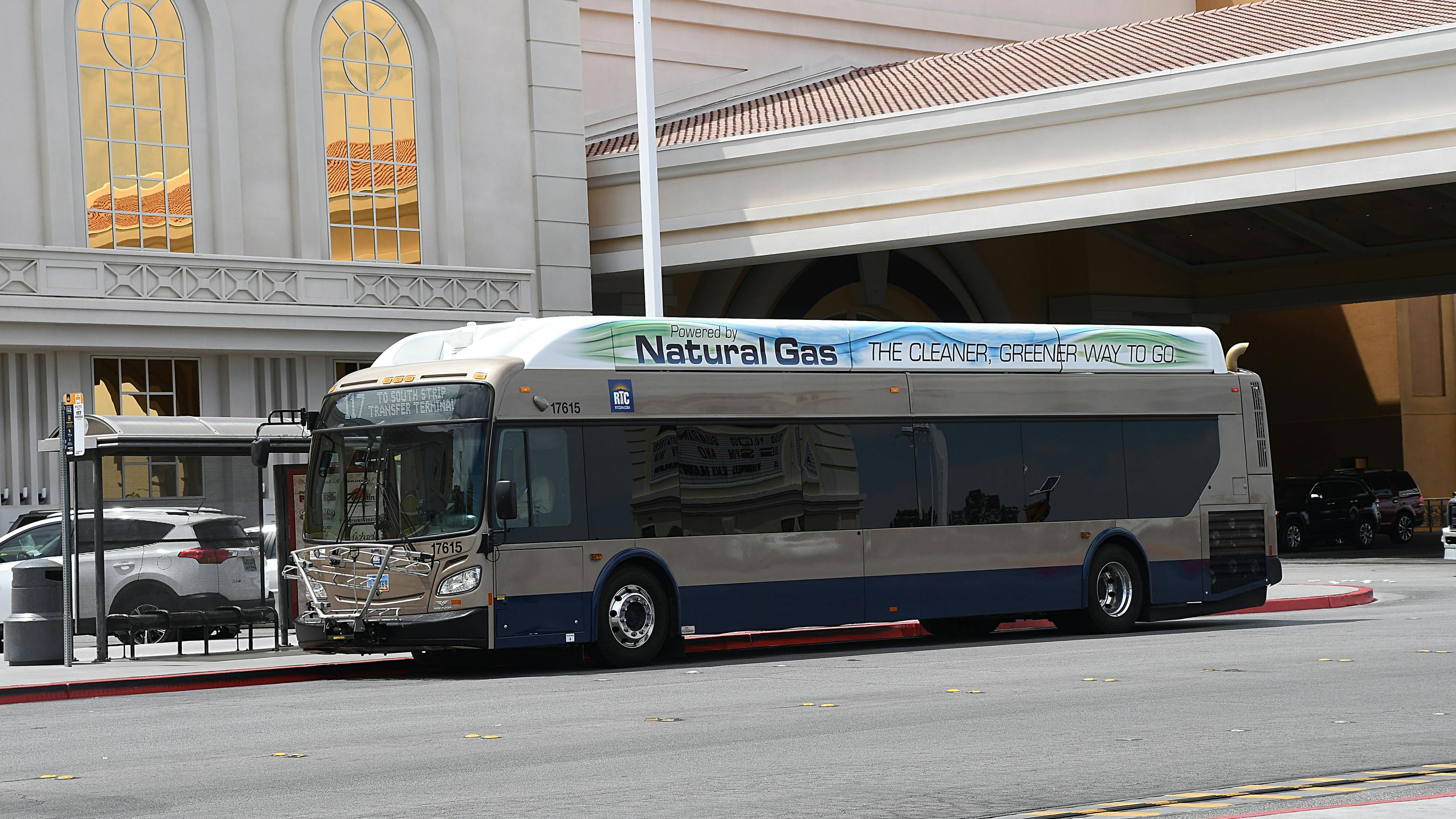 An RTC of Southern Nevada bus, unit 17615, in tan and navy livery with &ldquo;Powered by Natural Gas: The cleaner, greener way to go&rdquo; on its roof panel, displays &ldquo;117 To South Strip Transfer Terminal&rdquo; while stopped at a curb beside a large resort building.