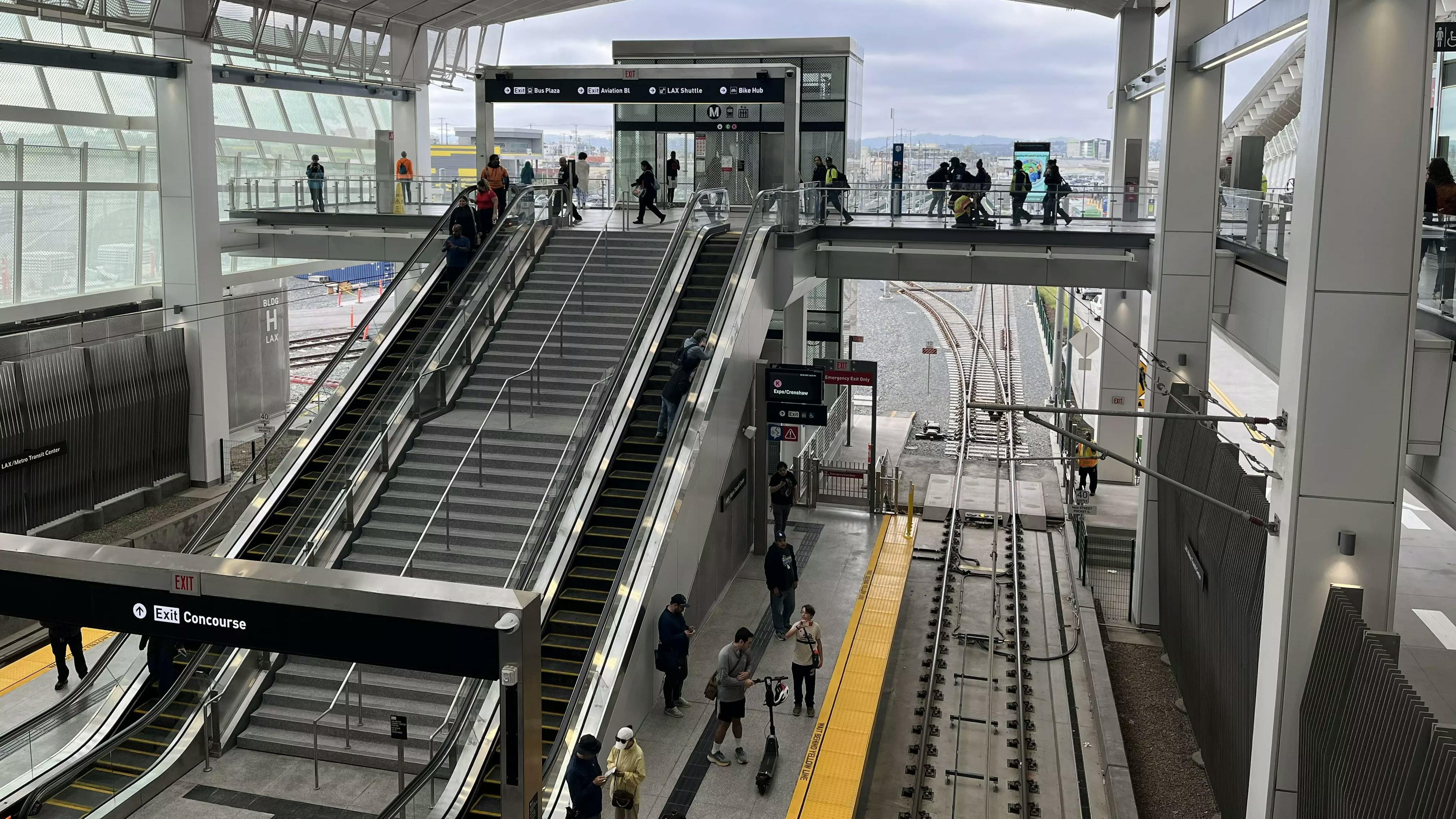 An elevated LAX/Metro Transit Center station, with escalators and stairs leading down to a light rail platform where riders wait, tracks visible below.