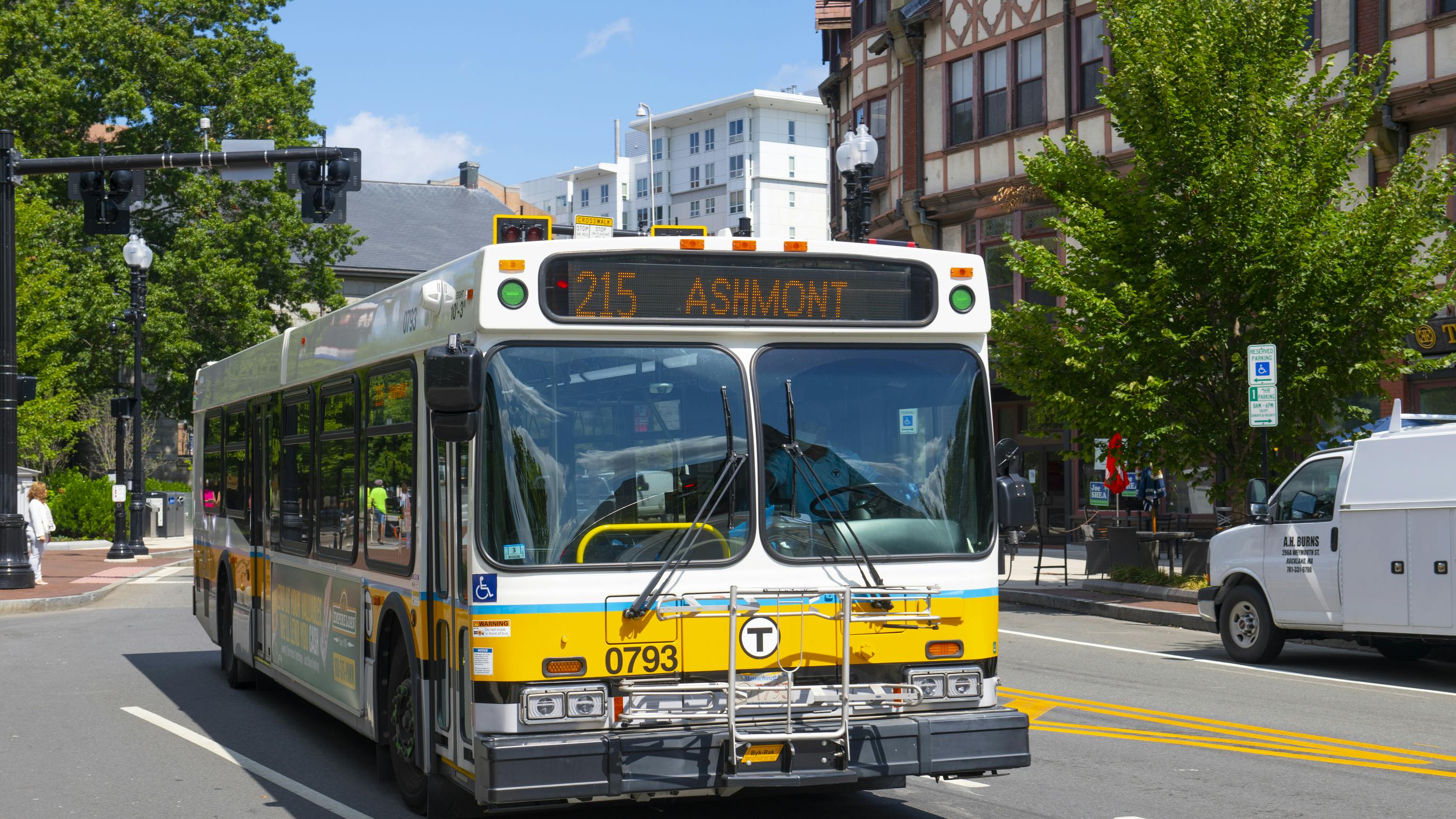 An MBTA bus, unit 0793, in white, yellow and blue livery displays &ldquo;215 Ashmont&rdquo; on its destination sign as it travels down a city street lined with trees and historic buildings, with a white work van at right.