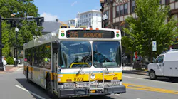 An MBTA bus, unit 0793, in white, yellow and blue livery displays “215 Ashmont” on its destination sign as it travels down a city street lined with trees and historic buildings, with a white work van at right.