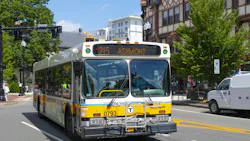 An MBTA bus, unit 0793, in white, yellow and blue livery displays “215 Ashmont” on its destination sign as it travels down a city street lined with trees and historic buildings, with a white work van at right.
