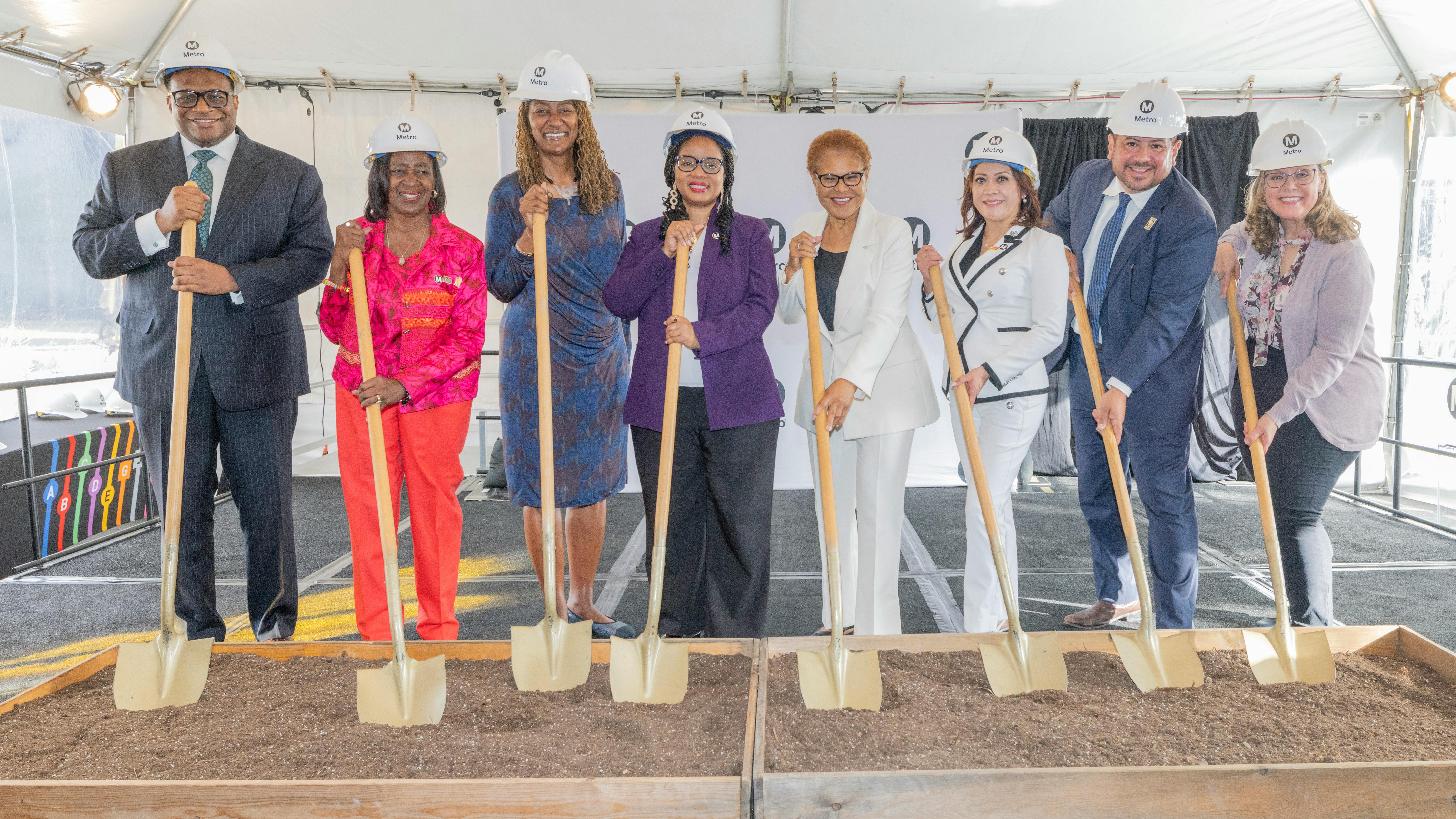 Eight officials in white Metro hard hats stand in a row holding gold-colored ceremonial shovels planted in two wooden boxes of soil, under a white tent with a Metro-branded backdrop.