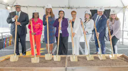 Eight officials in white Metro hard hats stand in a row holding gold-colored ceremonial shovels planted in two wooden boxes of soil, under a white tent with a Metro-branded backdrop.