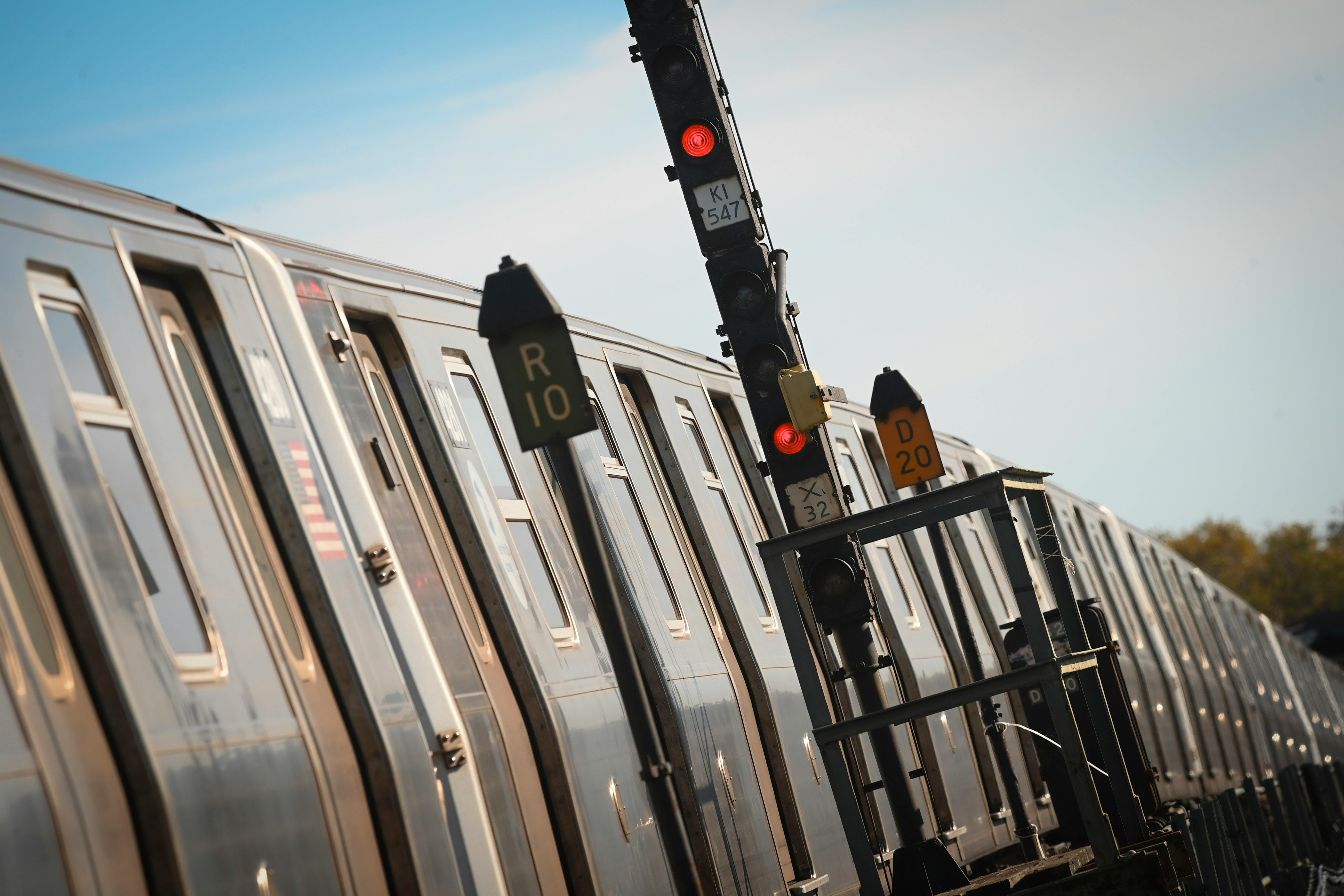 A tilted close-up of a stainless steel subway train stopped beside a wayside signal tower with red lights lit, route markers reading &ldquo;R10&rdquo; and &ldquo;D20&rdquo; and signal plates reading &ldquo;KI 547&rdquo; and &ldquo;X 32,&rdquo; against a blue sky.