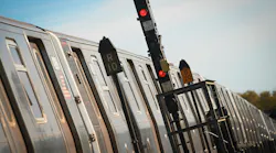 A tilted close-up of a stainless steel subway train stopped beside a wayside signal tower with red lights lit, route markers reading “R10” and “D20” and signal plates reading “KI 547” and “X 32,” against a blue sky.