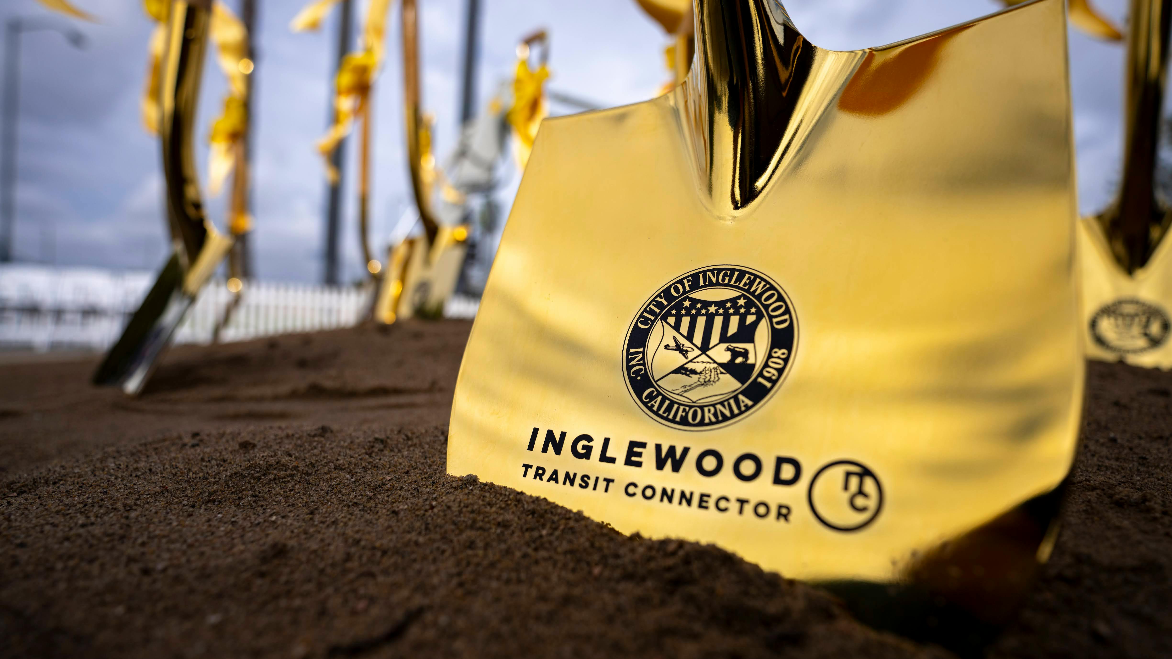 A close-up of a gold ceremonial shovel blade planted in soil, engraved with the City of Inglewood seal and the words &ldquo;Inglewood Transit Connector,&rdquo; with more gold shovels tied with gold ribbons blurred in the background.