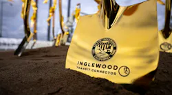 A close-up of a gold ceremonial shovel blade planted in soil, engraved with the City of Inglewood seal and the words “Inglewood Transit Connector,” with more gold shovels tied with gold ribbons blurred in the background.