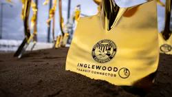 A close-up of a gold ceremonial shovel blade planted in soil, engraved with the City of Inglewood seal and the words “Inglewood Transit Connector,” with more gold shovels tied with gold ribbons blurred in the background.