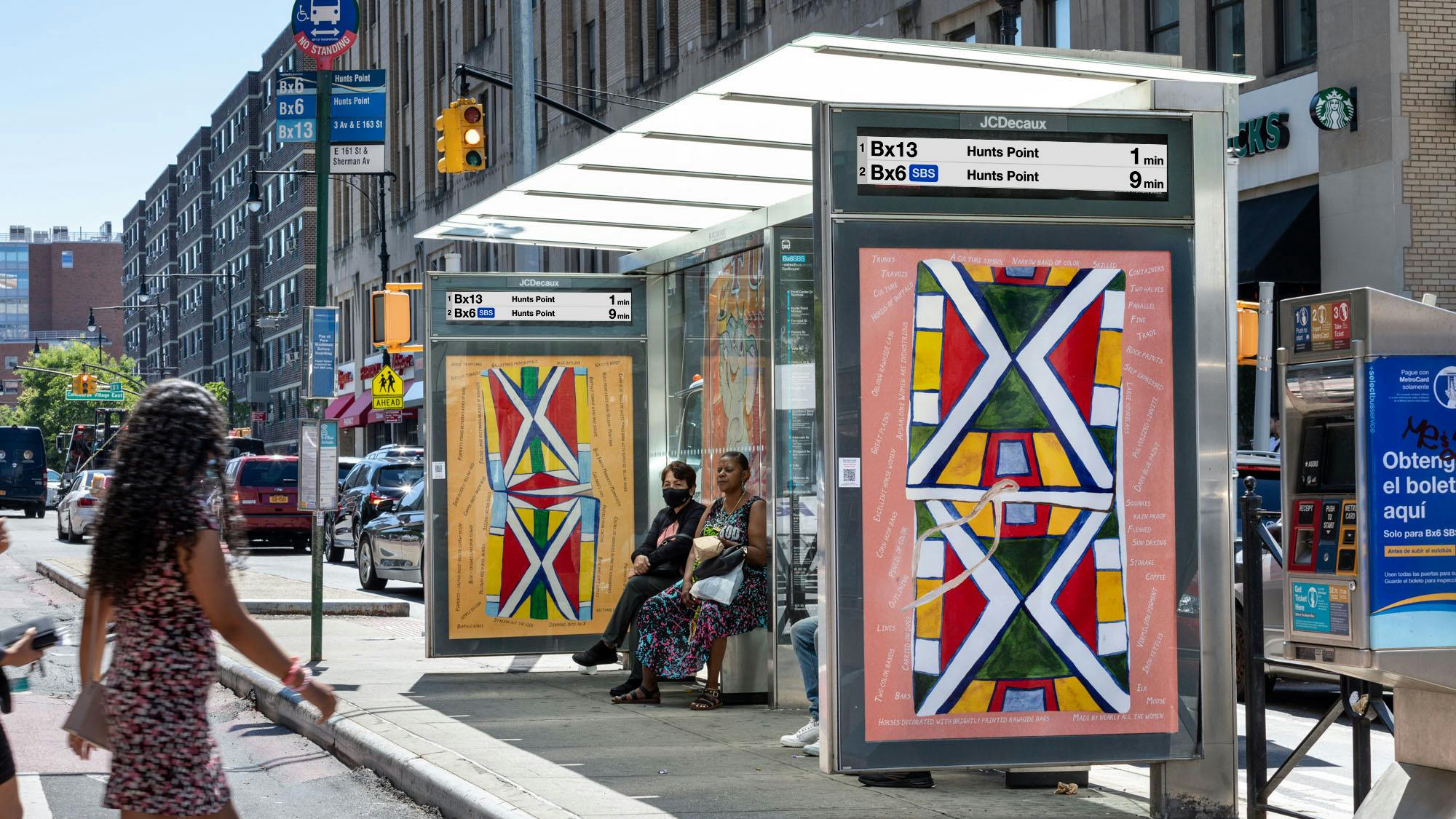 A JCDecaux bus shelter displaying colorful Indigenous-inspired artwork and a digital sign showing Bx13 and Bx6 SBS arrival times to Hunts Point, with riders seated on a Bronx street.