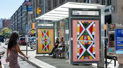 A JCDecaux bus shelter displaying colorful Indigenous-inspired artwork and a digital sign showing Bx13 and Bx6 SBS arrival times to Hunts Point, with riders seated on a Bronx street.