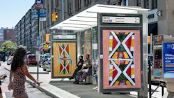 A JCDecaux bus shelter displaying colorful Indigenous-inspired artwork and a digital sign showing Bx13 and Bx6 SBS arrival times to Hunts Point, with riders seated on a Bronx street.
