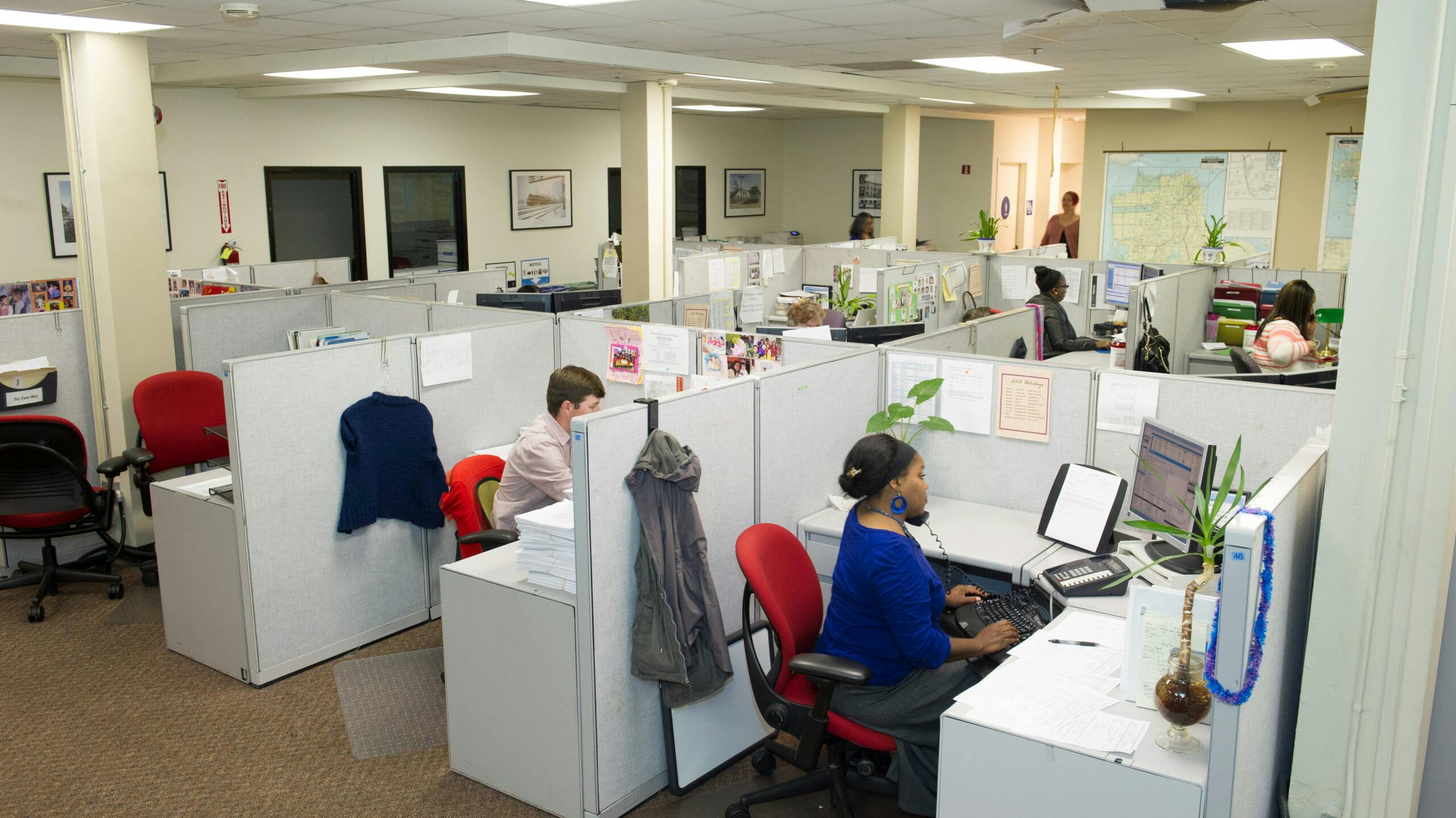 An overhead view of an open-plan call center with rows of gray cubicles, where employees at desks with computers and phones work, and a wall map hangs in the background.