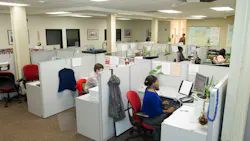 An overhead view of an open-plan call center with rows of gray cubicles, where employees at desks with computers and phones work, and a wall map hangs in the background.