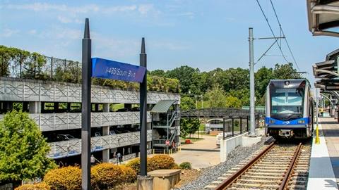 A light rail train approaches the I-485 South Blvd station platform, with a parking garage and trees in the background.