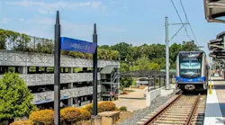 A light rail train approaches the I-485 South Blvd station platform, with a parking garage and trees in the background.