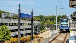 A light rail train approaches the I-485 South Blvd station platform, with a parking garage and trees in the background.