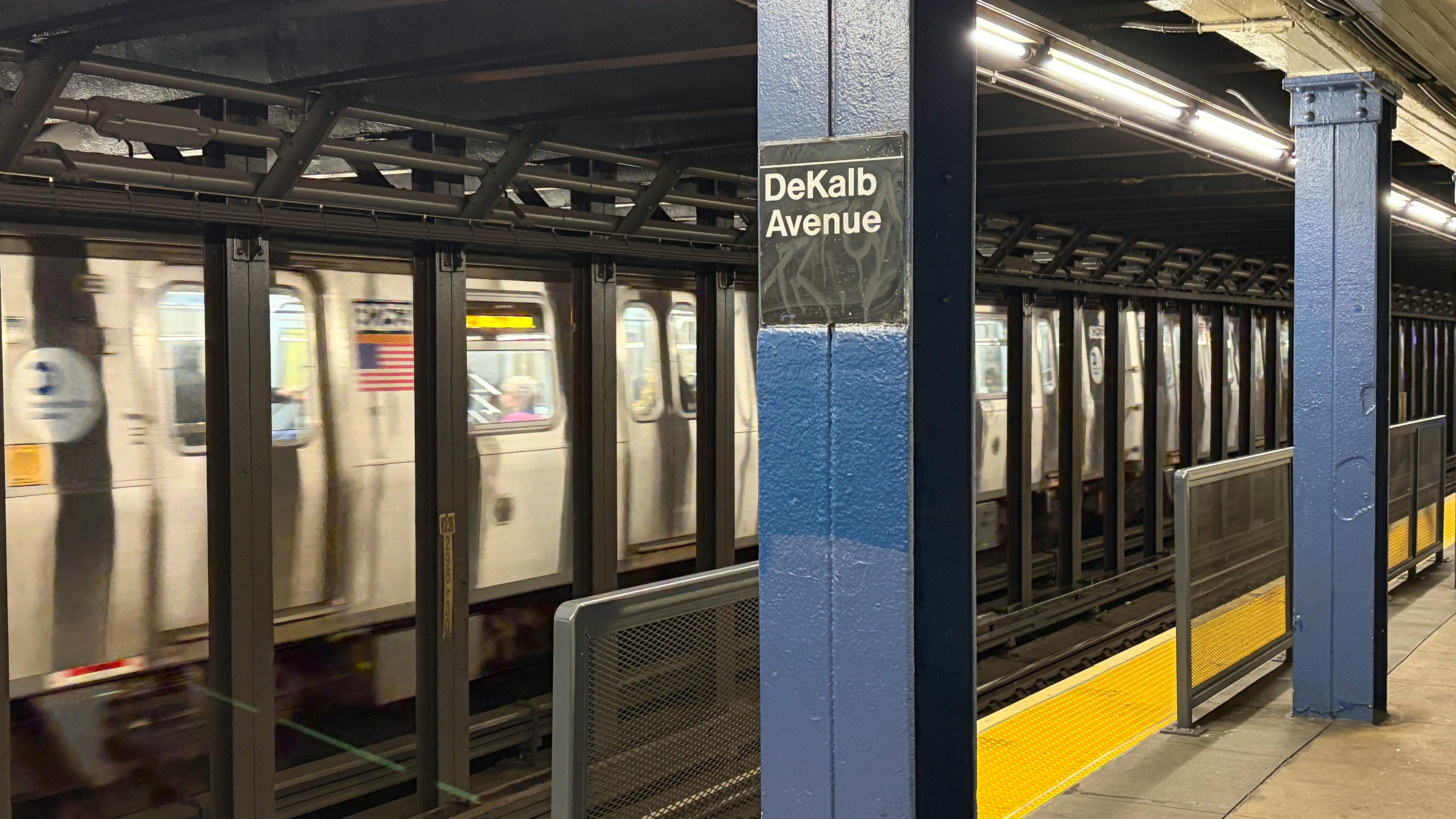 A subway train passes through the DeKalb Avenue station platform, with a blue column displaying the station name sign in the foreground.