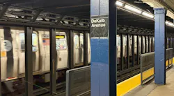 A subway train passes through the DeKalb Avenue station platform, with a blue column displaying the station name sign in the foreground.