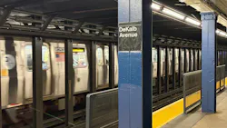 A subway train passes through the DeKalb Avenue station platform, with a blue column displaying the station name sign in the foreground.
