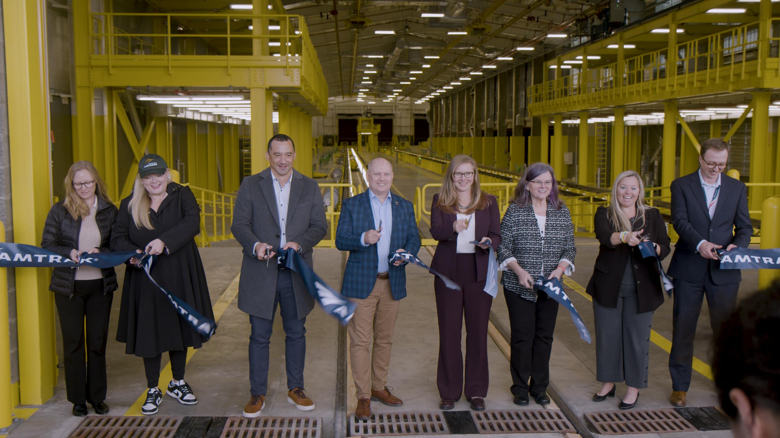 Eight people cut an Amtrak ribbon together inside a yellow-painted train maintenance facility in Seattle.