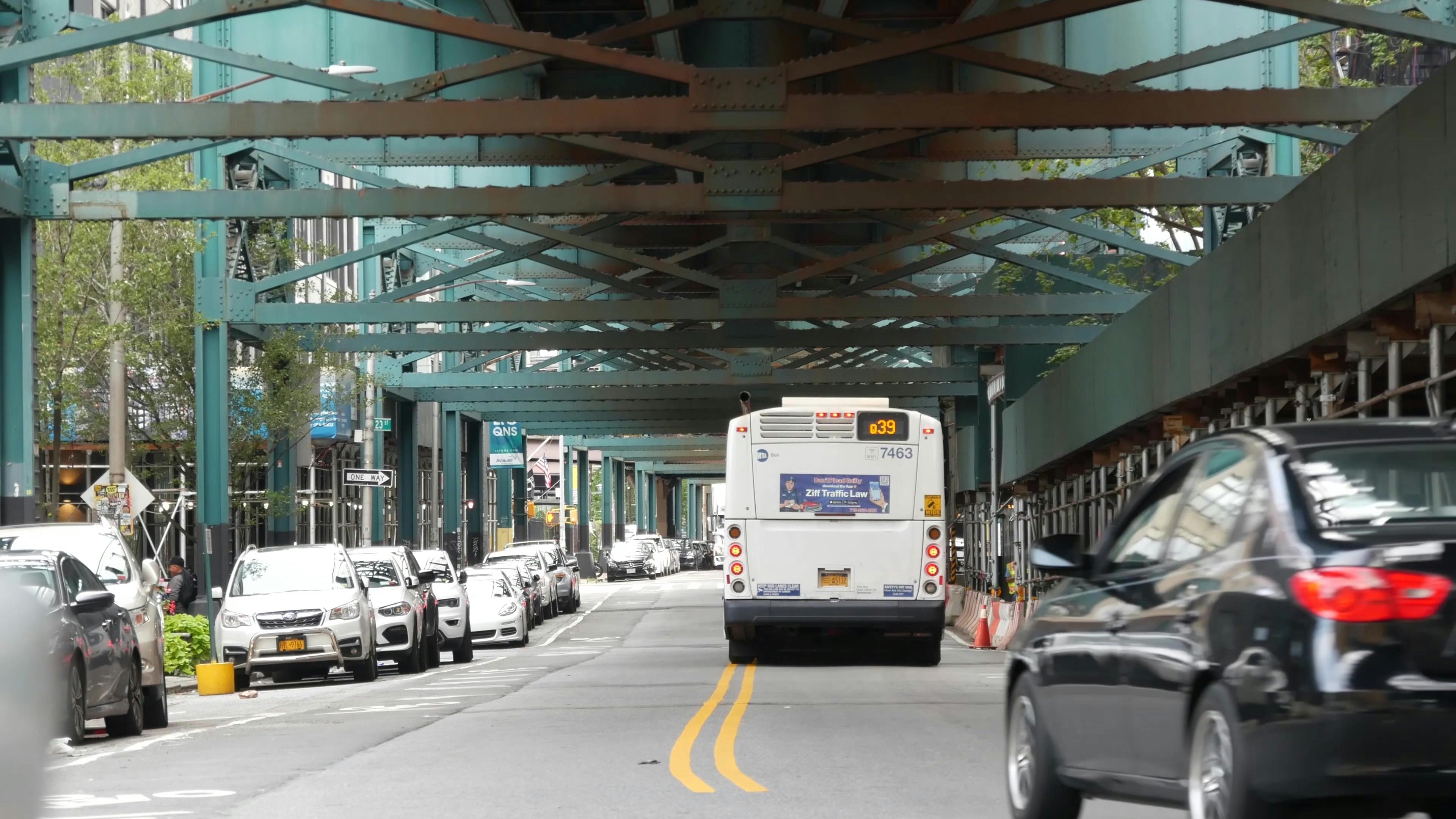 An MTA Q39 bus drives beneath an elevated subway structure on a city street, with parked cars lining both sides and a black sedan passing in the foreground.
