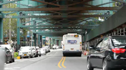 An MTA Q39 bus drives beneath an elevated subway structure on a city street, with parked cars lining both sides and a black sedan passing in the foreground.