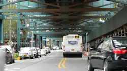 An MTA Q39 bus drives beneath an elevated subway structure on a city street, with parked cars lining both sides and a black sedan passing in the foreground.