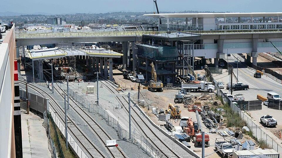 An elevated view of a rail station under construction, with several tracks in the foreground, a partly built station platform with canopy framing, an elevated guideway and station structure behind it, and excavators, trucks and other heavy equipment working across the site.