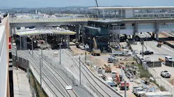 An elevated view of a rail station under construction, with several tracks in the foreground, a partly built station platform with canopy framing, an elevated guideway and station structure behind it, and excavators, trucks and other heavy equipment working across the site.