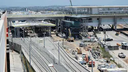 An elevated view of a rail station under construction, with several tracks in the foreground, a partly built station platform with canopy framing, an elevated guideway and station structure behind it, and excavators, trucks and other heavy equipment working across the site.
