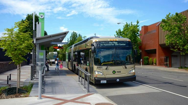 A tan and green articulated Vine bus, unit 6001, signed &ldquo;The Vine to Vancouver City Center,&rdquo; stops at a covered bus rapid transit station platform with a green pylon and real-time arrival display, as passengers wait on the sidewalk.