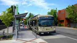 A tan and green articulated Vine bus, unit 6001, signed “The Vine to Vancouver City Center,” stops at a covered bus rapid transit station platform with a green pylon and real-time arrival display, as passengers wait on the sidewalk.