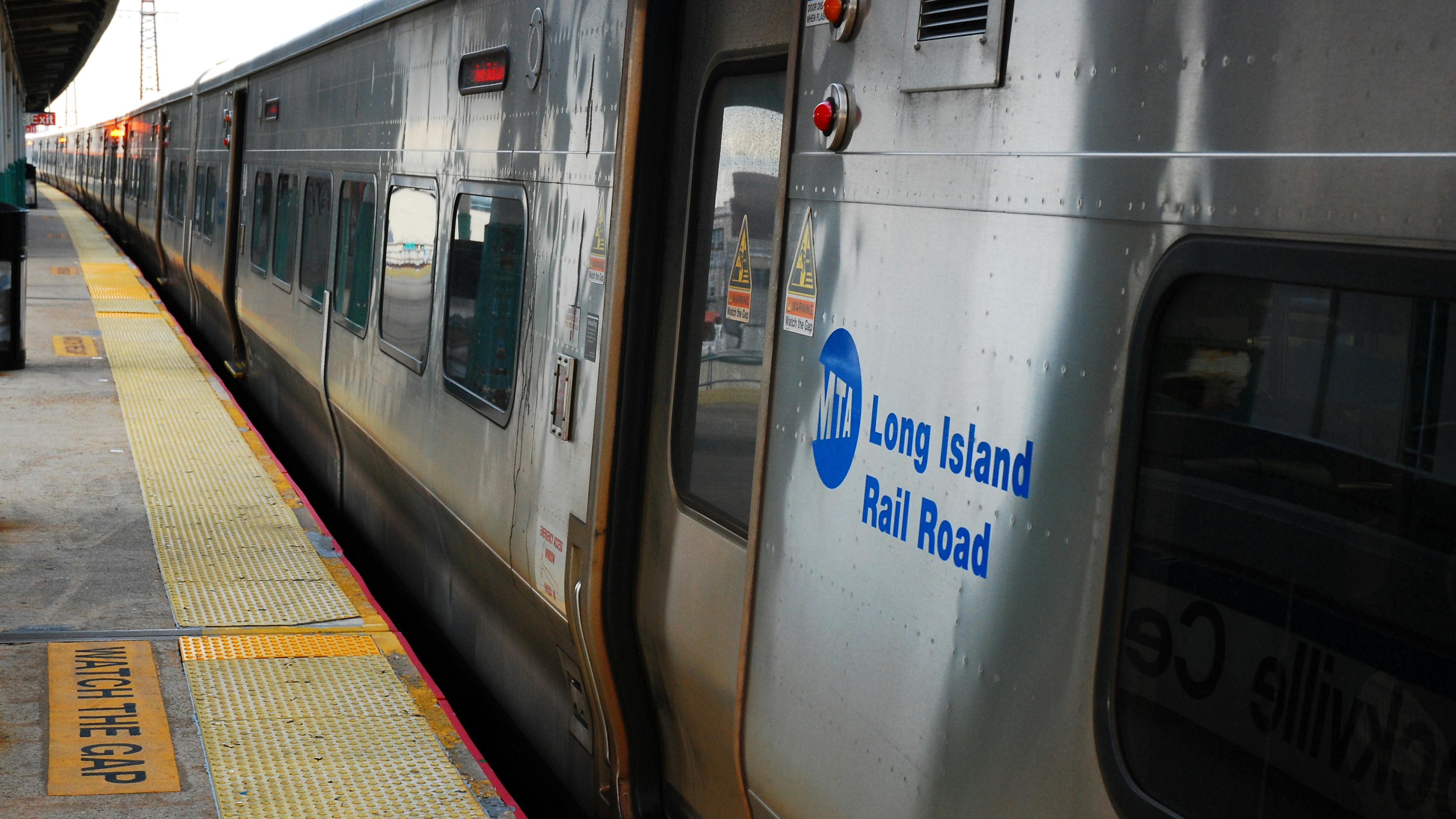 A Long Island Rail Road train stopped at a station platform, with its door open beside a &lsquo;Watch the Gap&rsquo; warning strip.