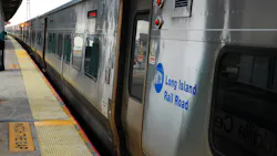 A Long Island Rail Road train stopped at a station platform, with its door open beside a ‘Watch the Gap’ warning strip.