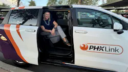 A smiling woman in a navy top and gray pants sits in the open side door of a white minivan with orange and purple graphics, a “PHX Link” logo on the door and “Powered by Via” on the rear window, parked in a lot with carports behind it.