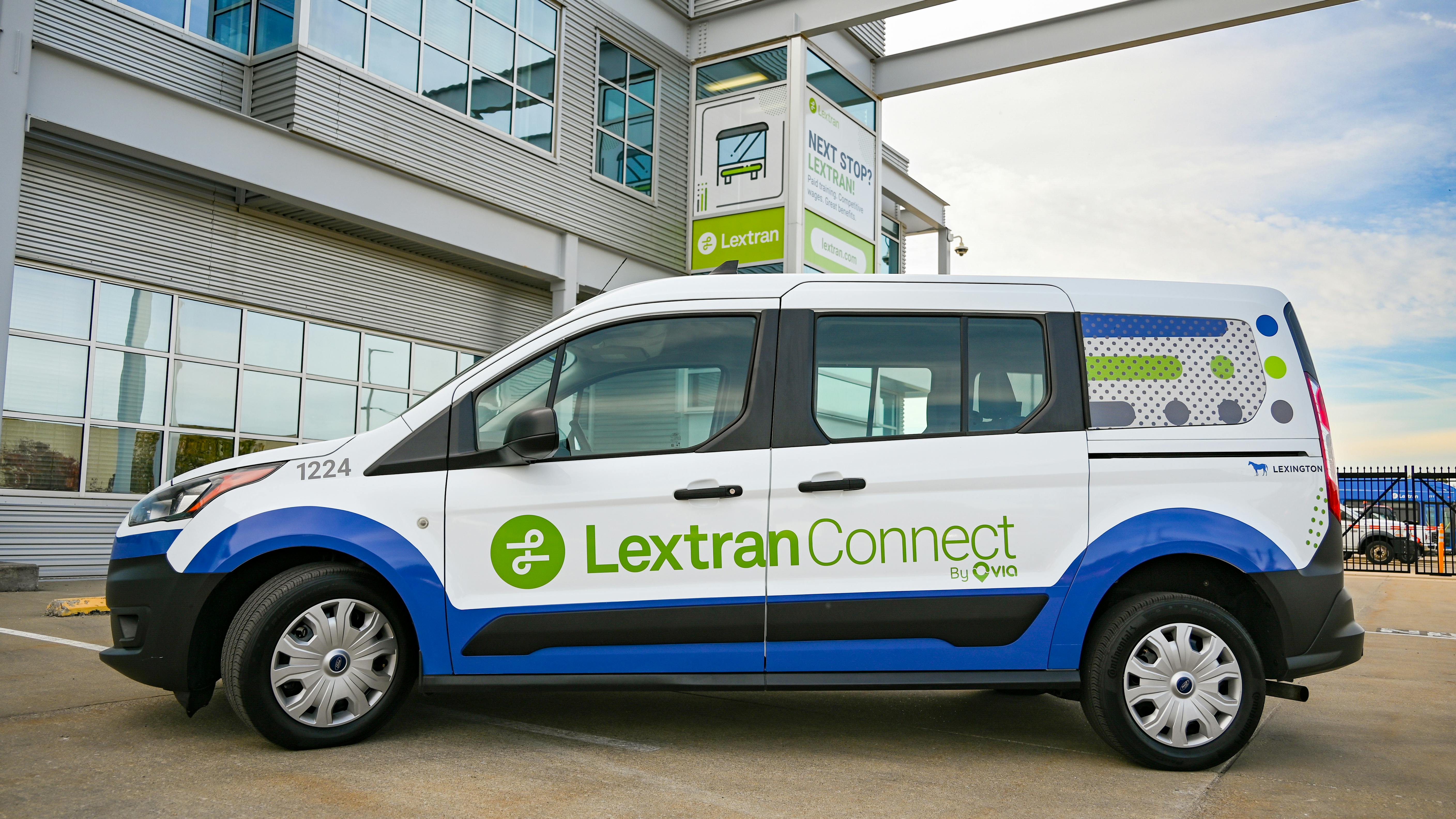 A white, blue and green Lextran Connect on-demand van, unit 1224, with &ldquo;By Via&rdquo; and &ldquo;Lexington&rdquo; branding, is parked in front of a gray Lextran facility with a recruiting sign reading &ldquo;Next stop? Lextran!&rdquo;