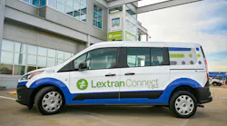 A white, blue and green Lextran Connect on-demand van, unit 1224, with “By Via” and “Lexington” branding, is parked in front of a gray Lextran facility with a recruiting sign reading “Next stop? Lextran!”